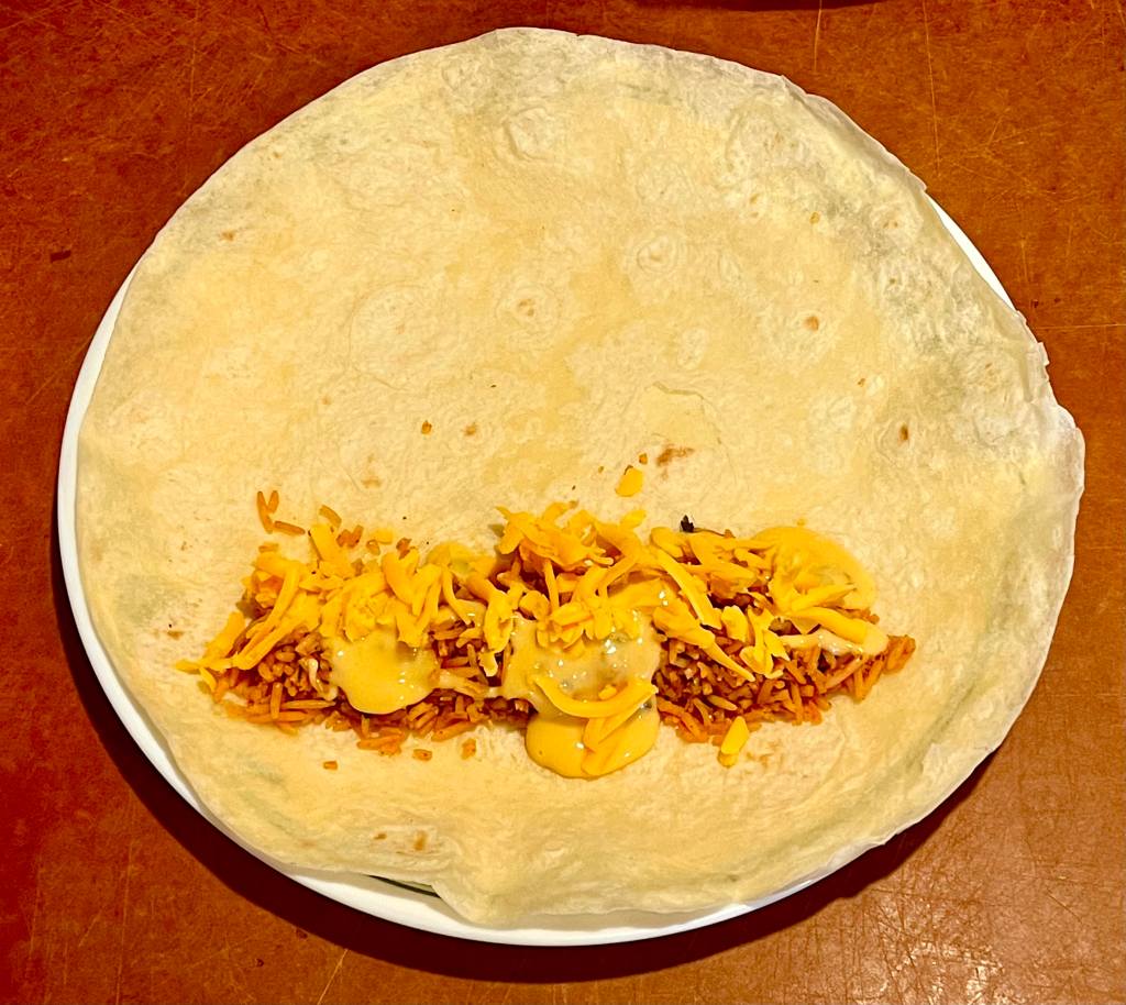 A steamed tortilla on a plate with rice and cheese filling for a bean burrito.