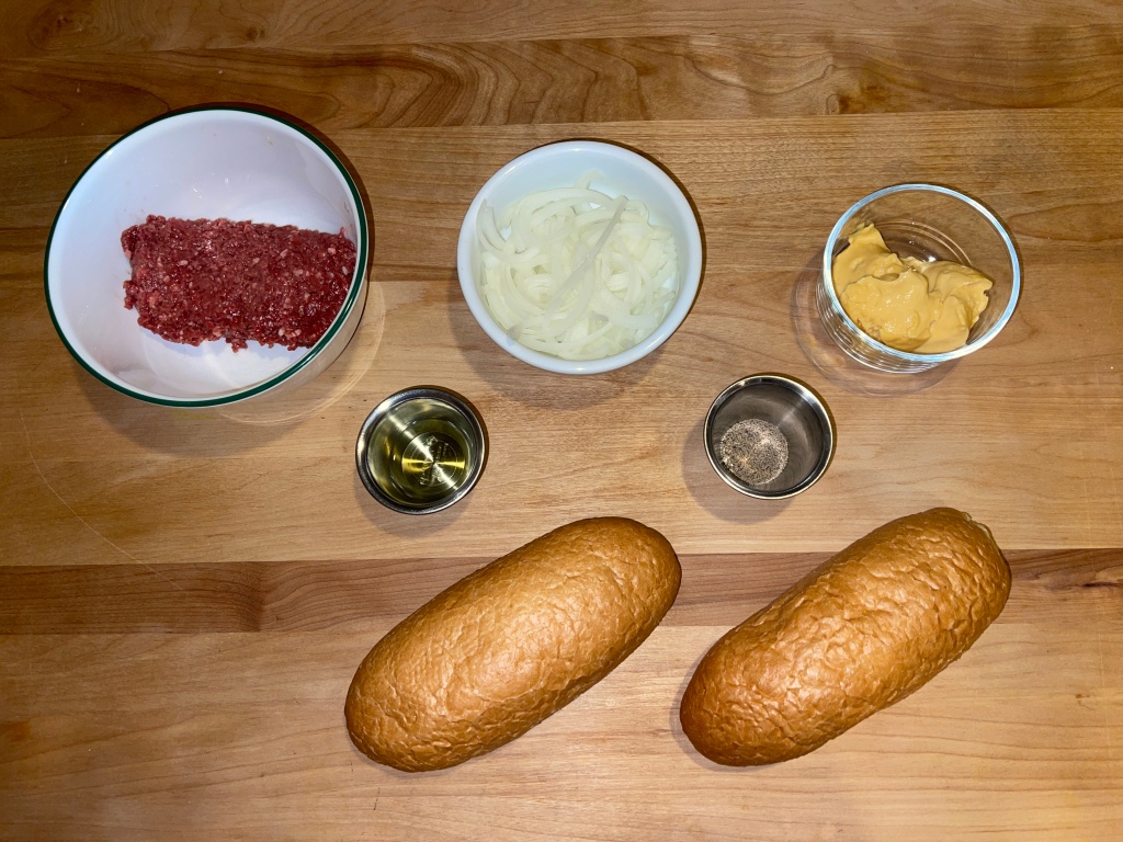 A photo showing the ingredients to make a veggie philly cheesesteak. This includes vegan beef, thinly sliced onions, cheese sauce, oil, black pepper, salt, and two long sandwich rolls.